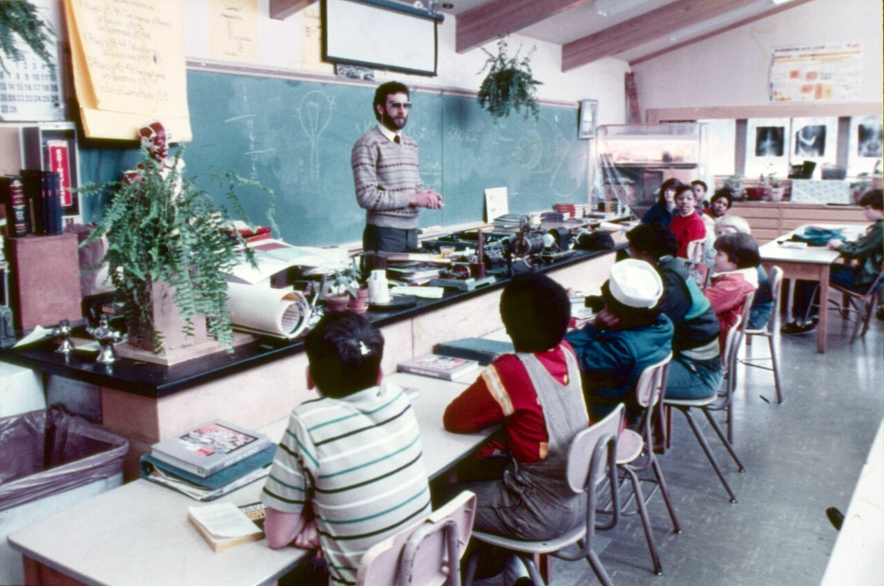 A teacher stands at a chalkboard while students look on in a public school classroom