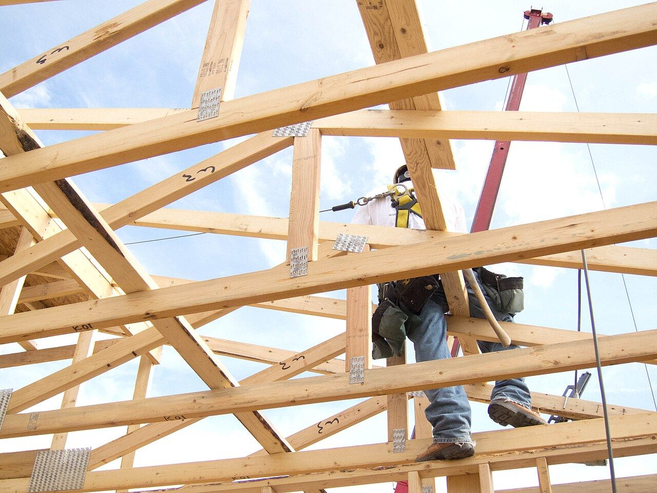 A construction worker wearing a safety harness walks across roof trusses on a house under construction