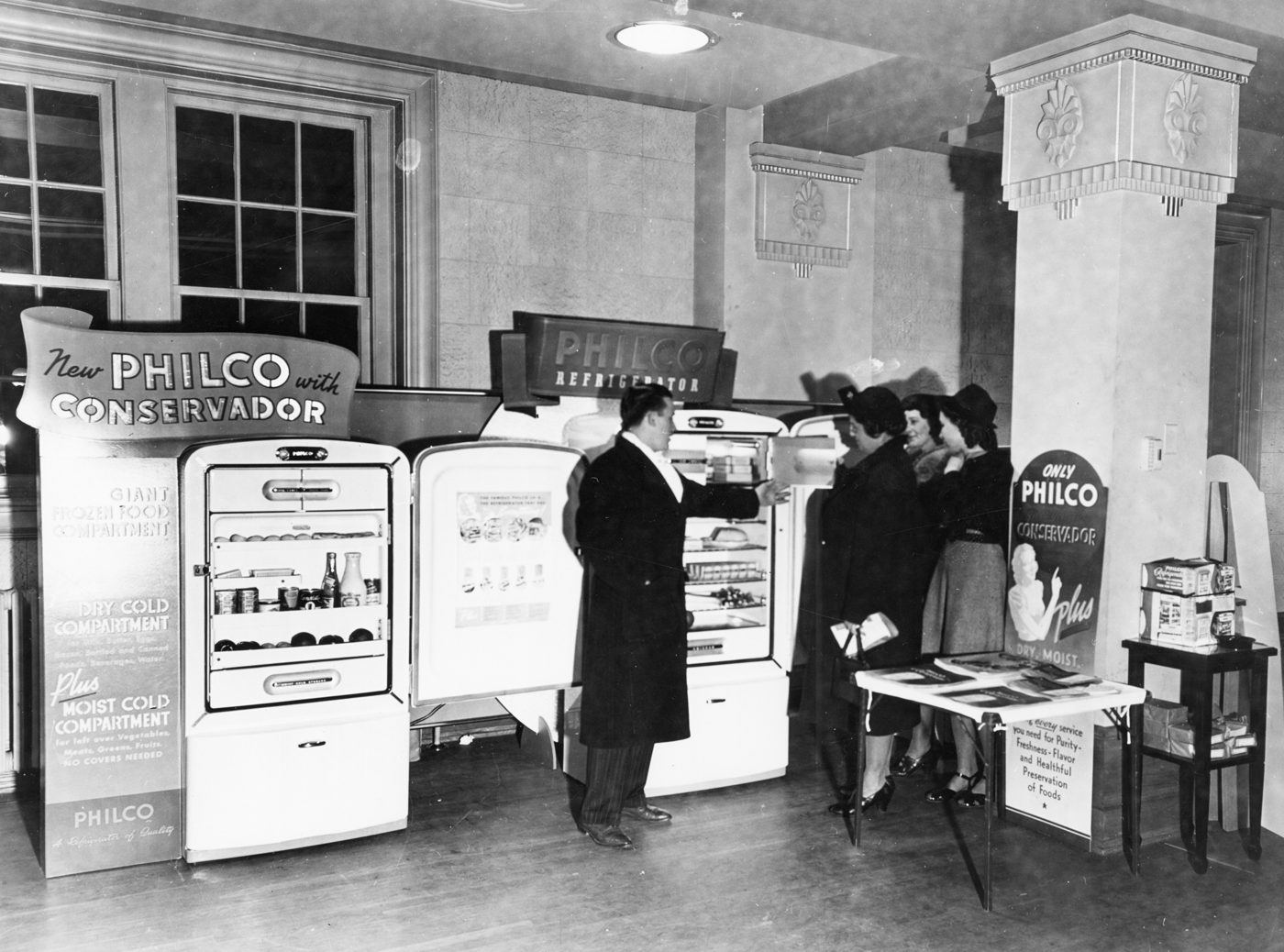 A 1940s appliance showroom with a salesman demonstrating refrigerators to customers